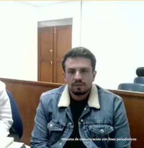 La imagen muestra a una persona sentada en un espacio interior que parece una sala institucional o de audiencias, debido a la presencia de mobiliario de madera y distribución tipo estrado.Primer plano:
En el centro de la imagen hay una persona sentada, mirando hacia la cámara. La persona tiene el cabello corto y lleva una chaqueta de mezclilla azul con cuello de tela clara (tipo borrego) sobre una camiseta oscura. La persona está ubicada detrás de una superficie o mesa de madera.
En el lado izquierdo de la imagen se alcanza a ver parcialmente otra persona vestida con una prenda blanca, aunque solo se observa una parte del hombro y brazo. En el lado derecho, al fondo, se distingue el respaldo de una silla de color oscuro.